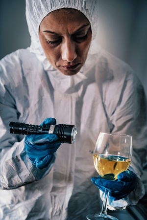 Police Crime Scene Investigator Examining Wine Glass with Forensic Light, Looking for Criminal Evidence.の写真素材