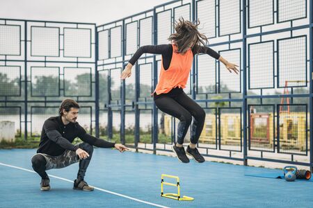 Hurdle jumping training outdoors.  Sporty woman jumping over the hurdle, fitness coach assistance.