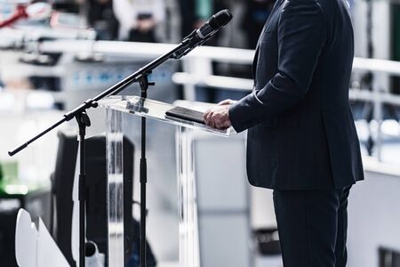 Male speaker wearing suit standing in front of microphones at media press conference.の写真素材