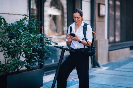 Businesswoman Standing and Using her Smart Phone with Electric Scooter next to her in The City.