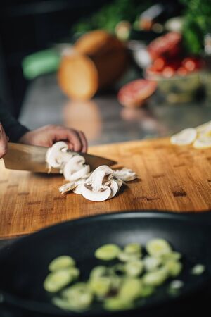 Cooking dinner. Chef holding a knife and cutting mushrooms on a wooden cutting board, close-upの写真素材
