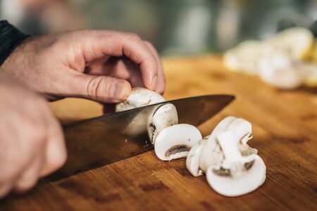 Cooking dinner. Chef holding a knife and cutting mushrooms on a wooden cutting board, close-upの写真素材