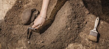 Archaeologist digging with hand trowel, recovering ancient pottery object from an archaeological site.の写真素材