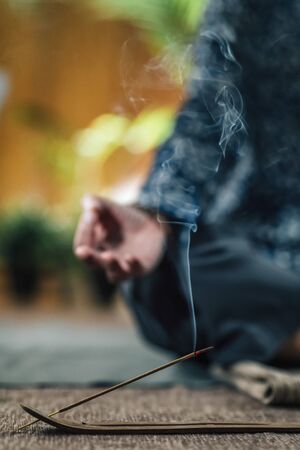Selective focus set on burning incense stick and smoke with woman meditating in background. Holding hands together at her chest in prayer.の写真素材