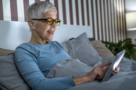 Mature woman wearing blue light blocking glasses with amber lenses, lying in bed before sleep,...