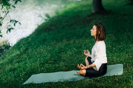 Young woman sitting in lotus position and practicing meditation near water in the natureの写真素材