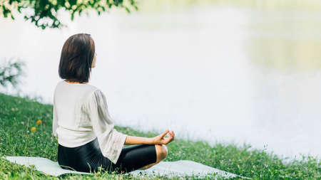 Young woman sitting in lotus position and practicing meditation near water in the natureの写真素材