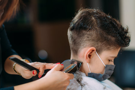 Hairdresser cutting boy's hair in hair salon, wearing protective mask