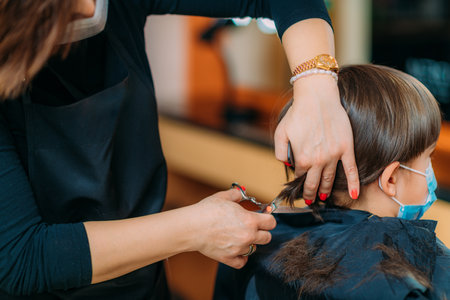Hairdresser cutting boy's hair in hair salon, wearing protective maskの写真素材