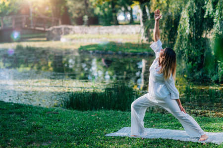 Yoga in nature, young woman in white, peaceful warrior pose, soft green nature background.の写真素材