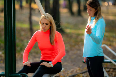 Female Friends Exercising in Public Park. Woman Doing Exercise for Legs on Machine. Autumn, Fall.