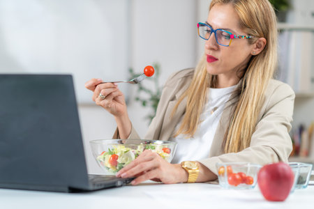 Healthy eating at work. Woman indulges in a delightful fresh salad at her workplace. With every bite, she embraces the nourishing flavors and invigorating energy of her nutritious meal, fueling her day with a refreshing and health-conscious choiceの写真素材