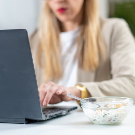 The perfect balance of work and wellness as a woman embraces healthy eating at her workplace. With a bowl of nutritious muesli and yogurt by her side, she savors each bite while efficiently typing on her laptop, optimizing her productivityの写真素材