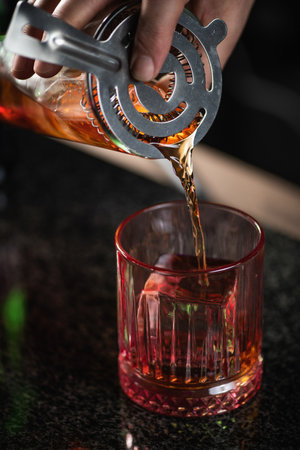 Skilled bartender pours Negroni cocktail through a cocktail drink strainer.