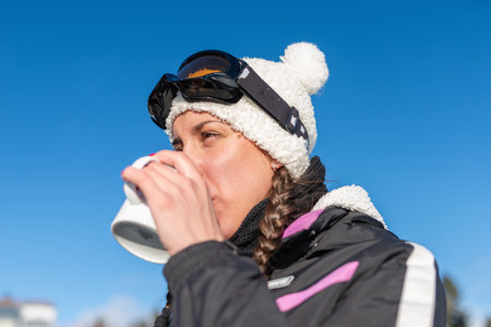 A woman enjoying a winter day on the mountain, sipping tea and embracing the snowy landscape. She is...