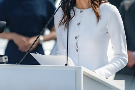 Female speaker conveys key updates in a close-up at a press conference, ensuring media clarity. Her...