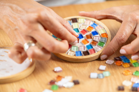 Arranging small craft pieces and gluing them onto coasters in a home studio. Captures precision,...