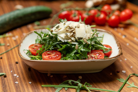 Fresh arugula salad with cherry tomatoes, shaved parmesan cheese and sunflower seeds, served in a ceramic dish on a rustic wooden table with fresh vegetables in the backgroundの写真素材