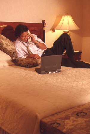 Male executive in hotel room talking on phone, with laptop computerの写真素材