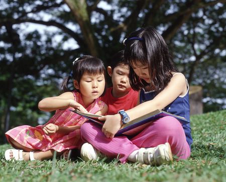 Three children sitting on grass reading a bookの写真素材