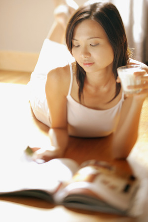 Young woman lying on floor holding a glass, flipping through magazineの写真素材