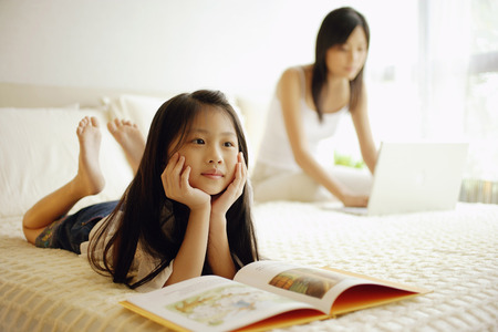 Mother and daughter in bedroom, mother using laptop, daughter lying down with book open in front of herの写真素材