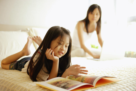 Mother using laptop, daughter flipping through bookの写真素材
