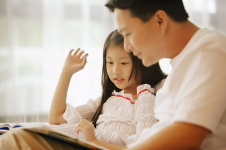 Father and daughter, sitting side by side, looking at bookの写真素材