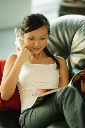 Young woman on sofa, holding phone and reading magazineの写真素材