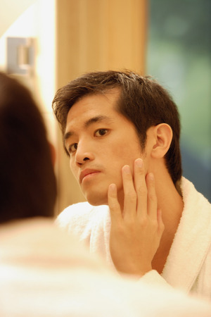 Young man touching his face, looking in mirrorの写真素材
