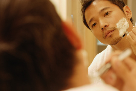Young man shaving, looking in mirror, over the shoulder viewの写真素材