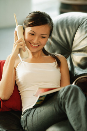 Young woman on sofa, holding phone and reading magazineの写真素材