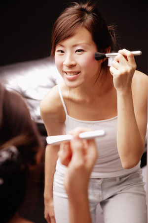 Young woman looking at mirror applying blusher with brushの写真素材