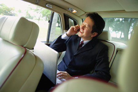 Businessman in backseat of car, using laptop and mobile phoneの写真素材