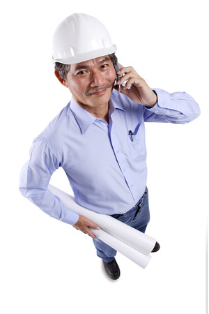 Mature man wearing construction hat, using mobile phone, carrying blueprints, looking at cameraの写真素材