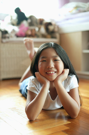 Girl in bedroom, lying on floor, hands on chin, looking at cameraの写真素材