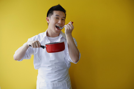Man wearing apron and holding saucepan, eating from spoonの写真素材