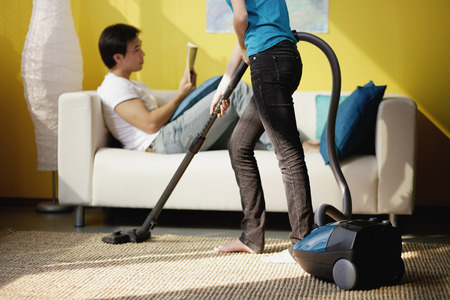 Woman vacuuming, man sitting on sofa reading magazineの写真素材