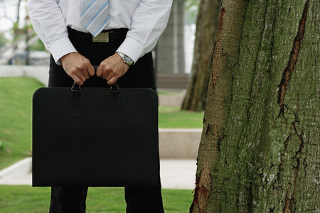 Businessman standing in park, holding briefcase, cropped imageの写真素材