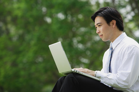 Businessman sitting in park with laptop, side viewの写真素材