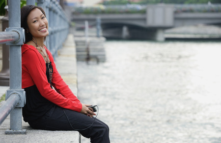 Woman sitting by river, listening to MP3 player, smiling at cameraの写真素材