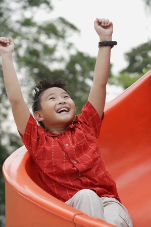 Boy coming down playground slide, arms outstretched, smiling, looking upの写真素材