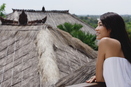 Young woman leaning on railing looking out at thatched roofsの写真素材