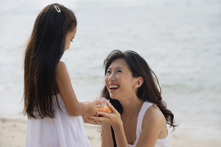Mother and daughter on beach, daughter handing conk shell to mother, smilingの写真素材
