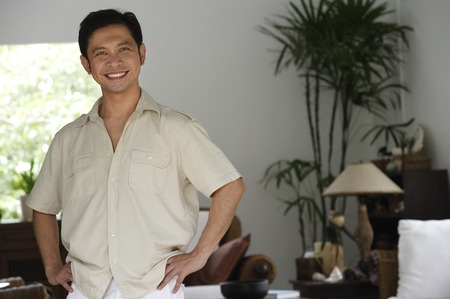 Man standing in living room, hands on hips, smilingの写真素材