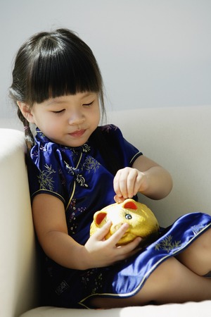 A small girl in blue silk cheongsam plays with a piggy bankの写真素材