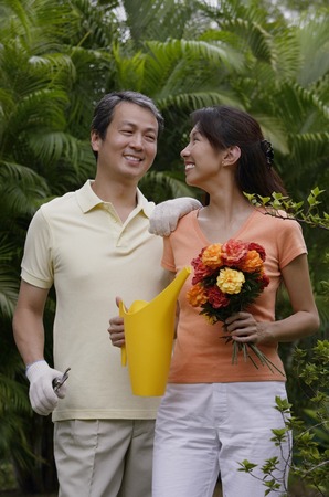 Couple in garden, woman holding bouquet of flowers and watering can, smiling at man next to herの写真素材