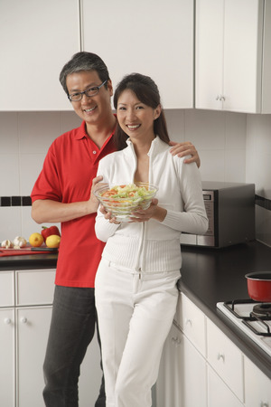 Man and woman in kitchen holding salad bowl and looking at camera smilingの写真素材