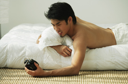 Young man lying in bed, holding alarm clockの写真素材