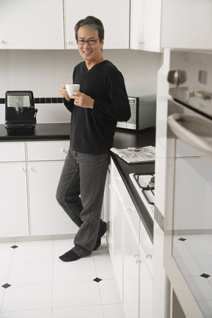 Man in kitchen, leaning on counter, holding coffee cup and smiling at cameraの写真素材